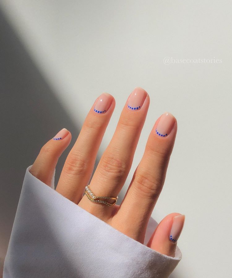 A imagem mostra uma mão com unhas decoradas. As unhas têm um esmalte base em tom claro e estão adornadas com pequenas bolinhas azuis formando uma linha curva próxima à ponta. O fundo é suave e neutro, destacando o design delicado e minimalista das unhas.
