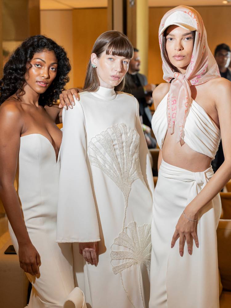 Foto de três mulheres com vestidos de noiva: uma de pele negra sem alças, outra de pele clara com manga comprida, e outra de pele clara com cropped, saia e lenço
