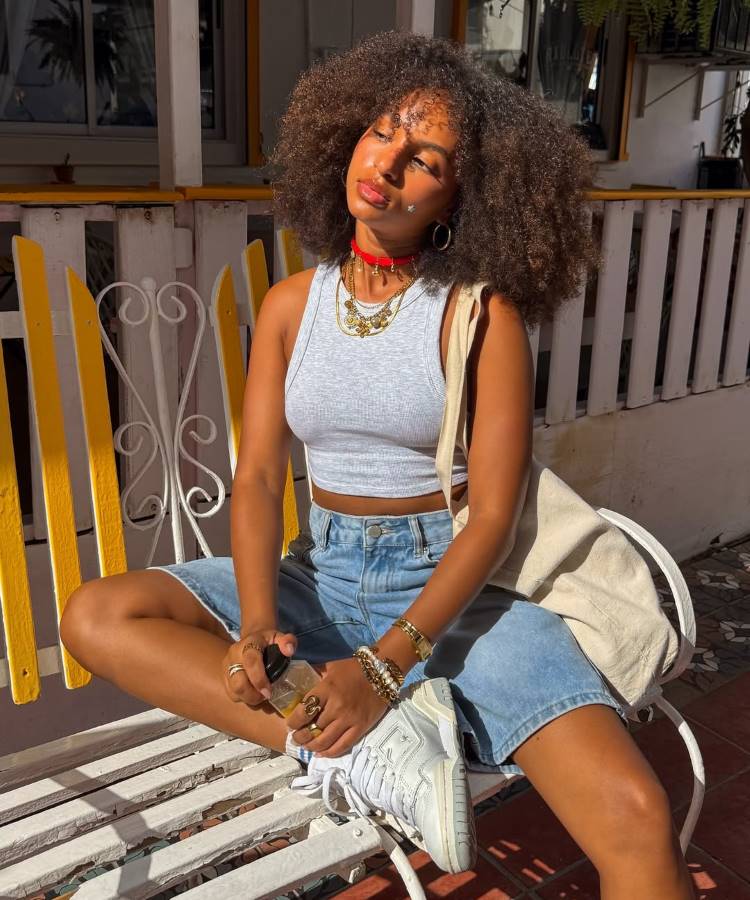 Uma jovem com cabelos cacheados e volumosos sentada em um banco de madeira. Ela usa um top cinza, jorts, tênis brancos e um colar vermelho, com a luz do sol batendo em seu rosto.