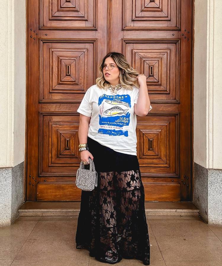 Uma mulher com cabelos loiros e óculos, vestindo uma camiseta branca com estampa de peixe e uma saia longa preta de renda. Ela segura uma bolsa prateada brilhante e posa em frente a uma grande porta de madeira esculpida.