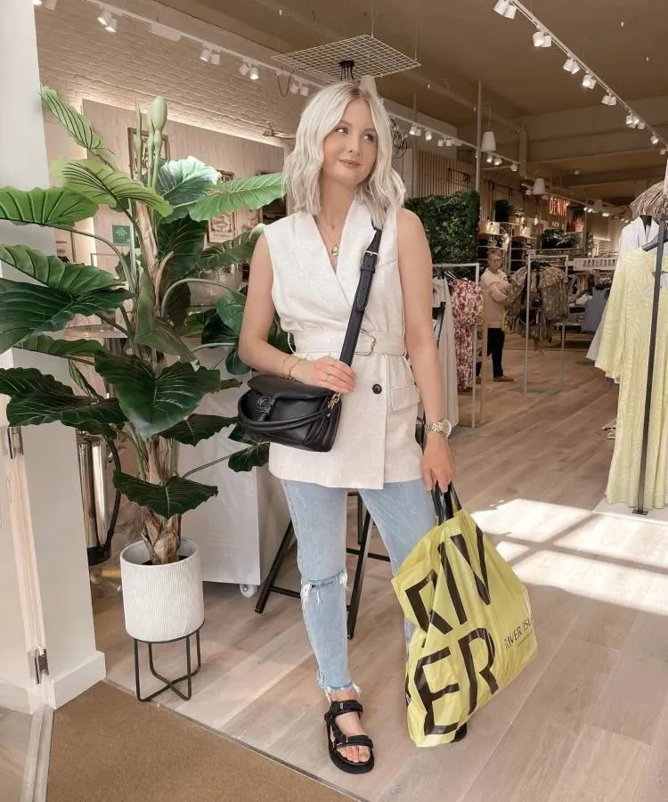 Uma mulher de cabelo platinado posa em uma loja usando um colete creme, calça jeans e uma bolsa combinando como sapato preto