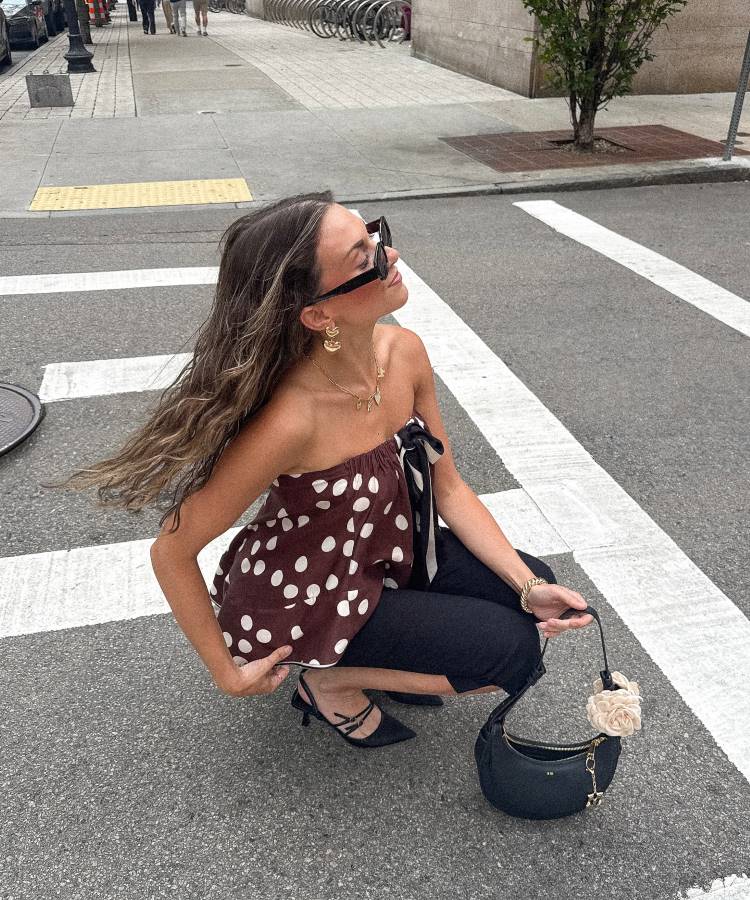 Mulher agachada em uma rua, vestindo uma blusa de bolinhas, calça preta e **sapato preto** de salto agulha. Ela usa óculos escuros, joias douradas, e segura uma bolsa preta. A foto é de corpo inteiro.