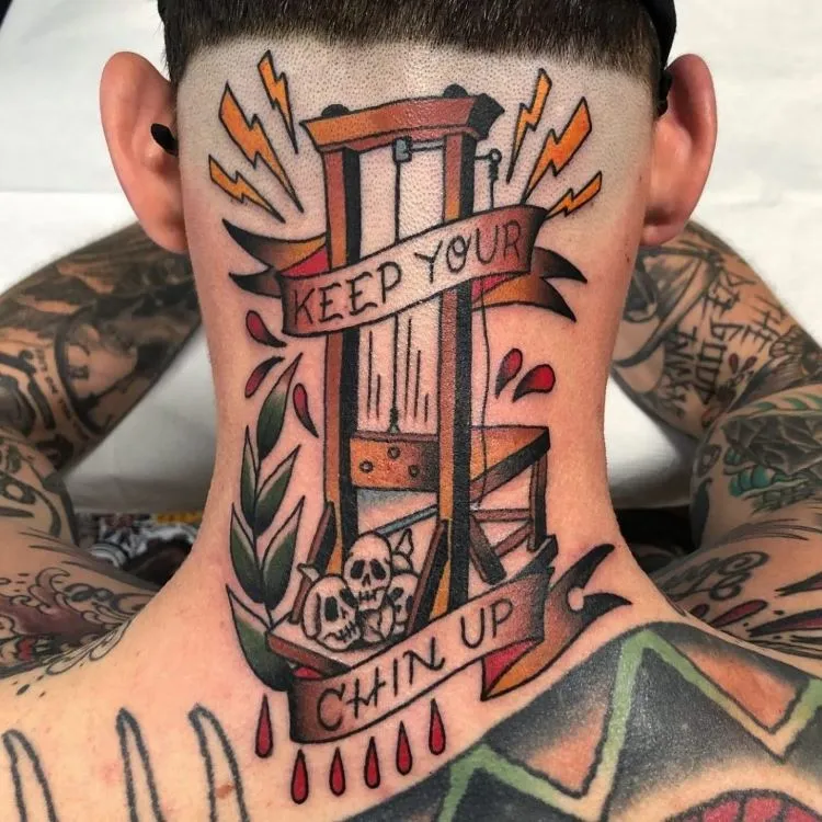 A imagem mostra uma pessoa de costas com cabelo curto. Ela tem uma tatuagem no pescoço de uma guilhotina com as palavras "Keep Your Chin Up" escritas em uma faixa. A tatuagem é colorida e detalhada, cobrindo a parte de trás do pescoço e parte das costas.