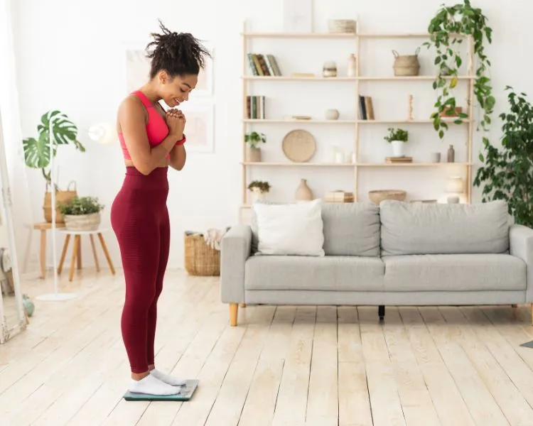 Mulher de pele escura usando roupa de fazer exercício em cima de balança na sala com sofá, estante decorada e plantas