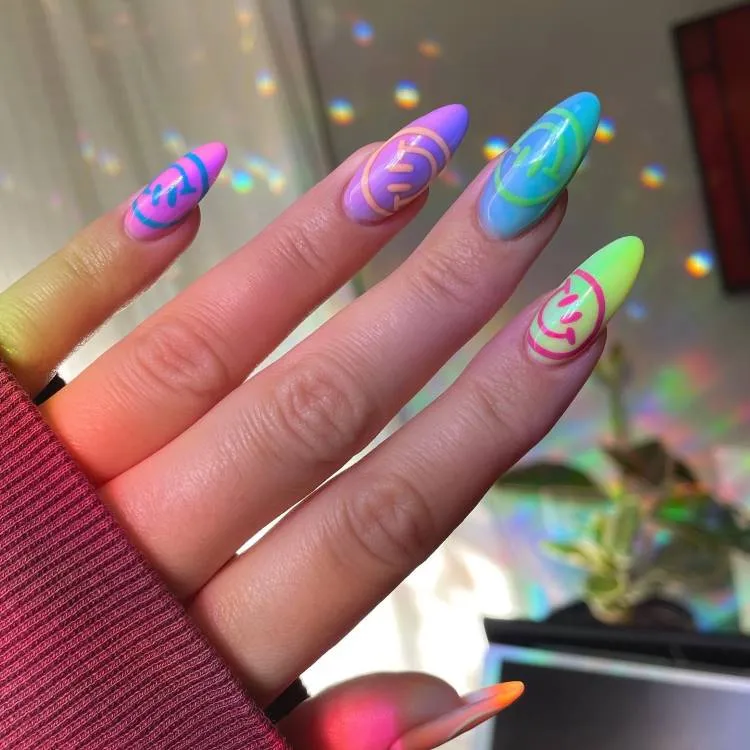A imagem mostra um close-up de uma mão com unhas longas e pontiagudas, pintadas em cores neon brilhantes. Cada unha tem uma combinação diferente de cores e padrões, incluindo redemoinhos e formas abstratas. As unhas exibem um efeito ombré, misturando uma cor na outra. O fundo mostra reflexos de luz criando padrões semelhantes a arco-íris nas superfícies ao redor da mão, sugerindo a presença de um objeto refletivo ou prismático próximo, que não é visível diretamente na imagem.

