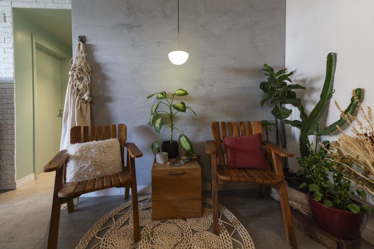 A imagem mostra um canto aconchegante com duas cadeiras de madeira, cada uma decorada com almofadas coloridas. Entre elas, há uma pequena mesa de madeira com uma planta em vaso. No chão, um tapete de crochê circular adiciona textura ao ambiente. Ao fundo, uma parede de cimento queimado destaca o espaço, enquanto várias plantas em vasos, incluindo um cacto, trazem um toque verde e natural. A iluminação pendente suave complementa a decoração. Este é um excelente exemplo de como decorar apartamento com plantas, criando um ambiente acolhedor e equilibrado, onde o verde das plantas contrasta com os tons neutros e terrosos da mobília e das paredes.