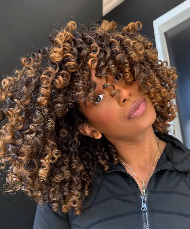 Uma mulher com um volumoso corte de cabelo cacheado, com cachos em tons de castanho e loiro, olha para a câmera. Ela usa um piercing no nariz e uma blusa preta com zíper.