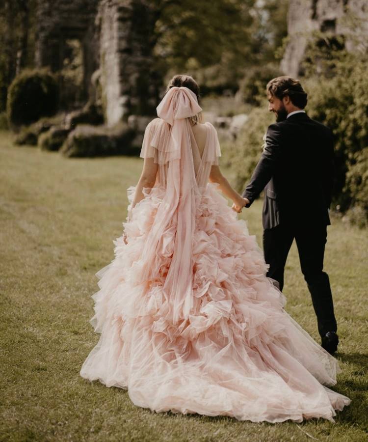 Uma noiva com um exuberante vestido rosa de babados e um grande laço nas costas, de mãos dadas com o noivo de terno escuro, caminha em um campo gramado com ruínas ao fundo.







