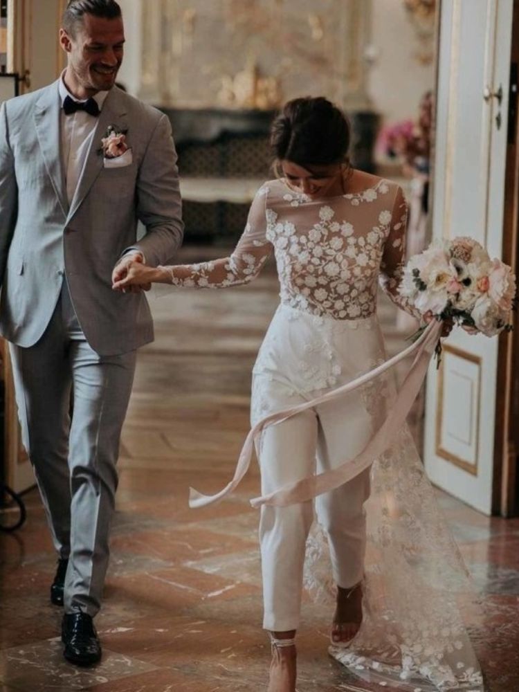 Mulher de pele clara usando blusa transparente com flores brancas bordadas e calça branca, segurando a mão de homem usando terno cinza
