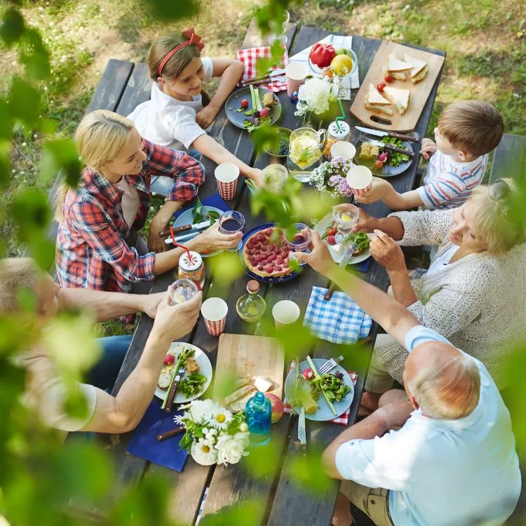 A imagem mostra uma família reunida ao redor de uma mesa de madeira ao ar livre, celebrando um Dia dos Pais criativo com um piquenique. Há alimentos como frutas e sanduíches, e todos parecem estar se servindo e desfrutando da companhia uns dos outros. As faces estão obscurecidas para privacidade.