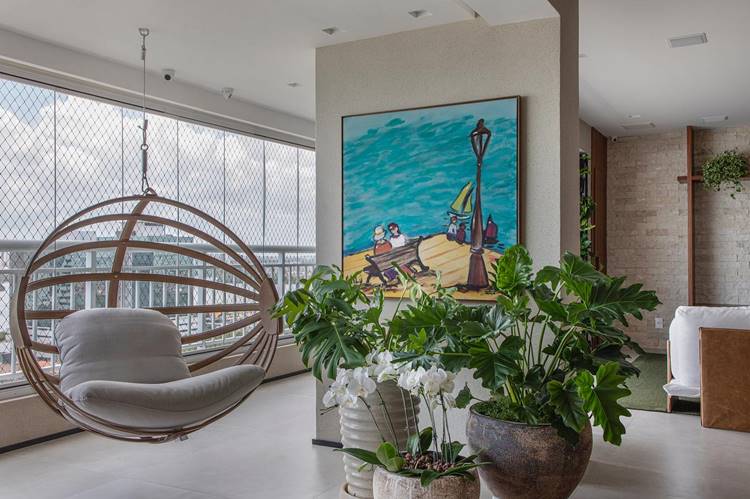 Decoração de verão de um interior moderno e elegante. Há uma cadeira suspensa circular com almofadas cinza clara na frente de janelas amplas que oferecem uma vista para o exterior. Uma grande pintura colorida está pendurada na parede à direita, retratando duas pessoas em um banco perto de um poste de luz, com o mar azul ao fundo. Várias plantas verdes estão dispostas pelo ambiente, incluindo um vaso grande no chão com flores brancas. A parede à direita tem uma textura de tijolos aparentes e há prateleiras embutidas contendo mais plantas. O ambiente é iluminado e arejado, com um piso claro e paredes brancas.


