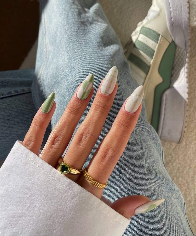 A imagem mostra unhas longas decoradas em tons de verde e branco, com designs geométricos, linhas xadrez e corações. A mão usa anéis dourados elegantes, incluindo um com pedra verde. O fundo exibe jeans e tênis verde pastel.