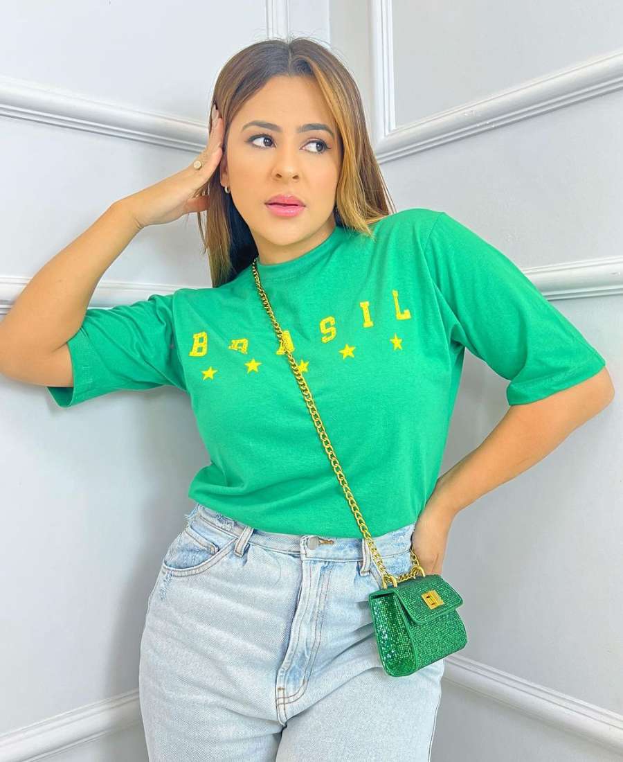 Imagem com fundo de cômodo de casa, com parede. No centro, mulher usando roupas para a copa do mundo: camiseta verde escrito de amarelo "Brasil", calça jeans e bolsa brilhante verde.