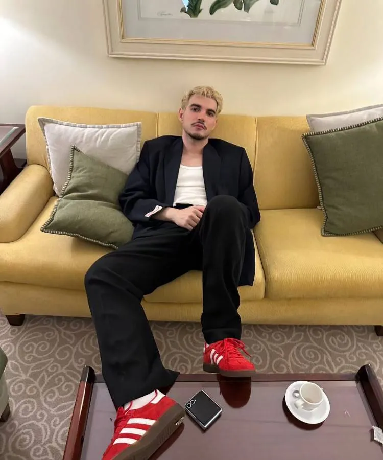 Um homem de cabelo loiro curto e barba bem aparada relaxa em um sofá amarelo. Usa blazer azul-marinho, camiseta branca, calças pretas e tênis vermelhos da Adidas. O ambiente é moderno, com mesa de centro e arte botânica na parede.
