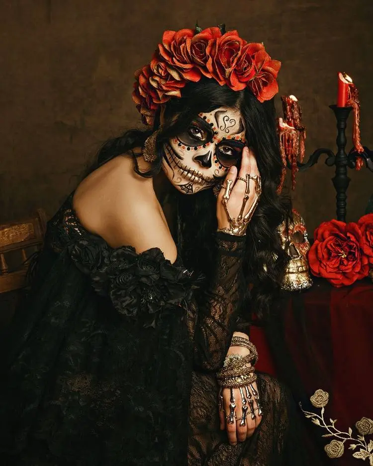 Mulher de pele clara usando maquiagem de caveira mexicana em preto e vermelho, vestido de renda preto e flores vermelhas no cabelo