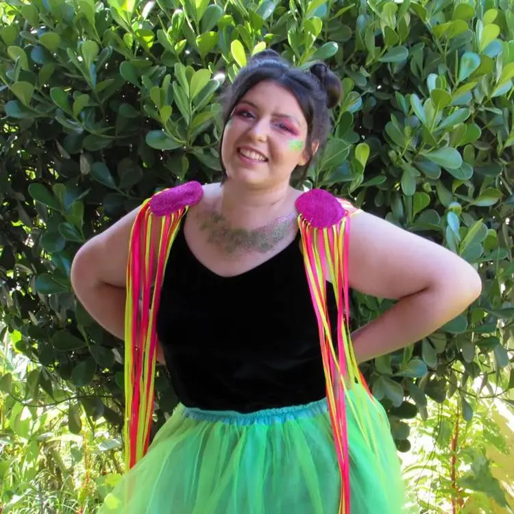 Mulher de pele clara com body preto, saia verde e ombreira de fitas rosas e amarelas 