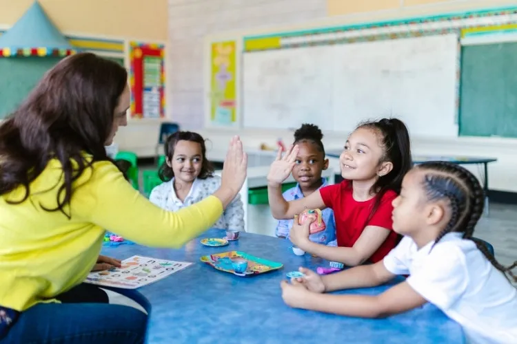 Professora em mesa com alunas, fazendo hi-five com uma delas