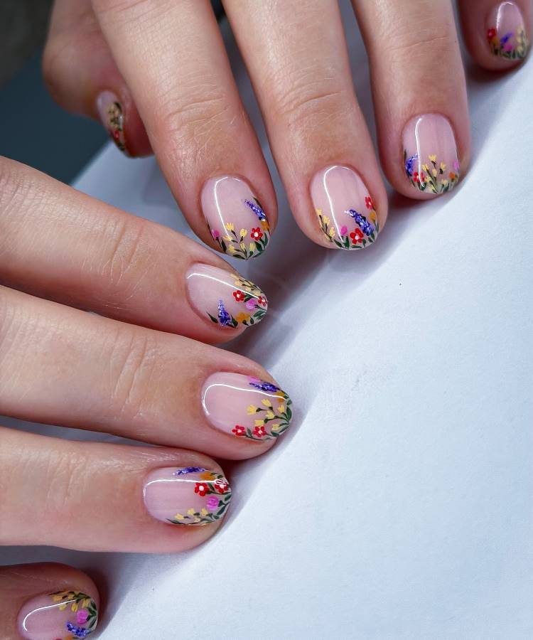 Unhas de gel decoradas com esmalte translúcido nude e detalhes florais coloridos, incluindo flores vermelhas, amarelas e lilases, cercadas por folhas verdes. O design delicado e elegante transmite um estilo primaveril sobre um acabamento suave e brilhante.