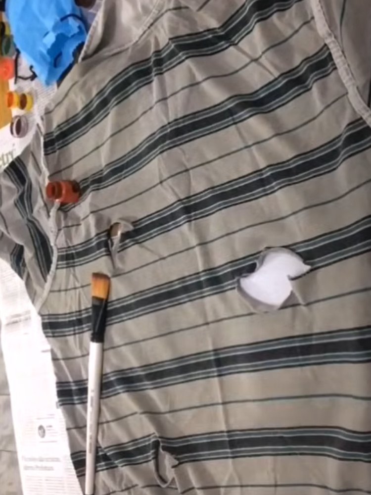 Mulher de pele clara segurando pincel, em frente à camisa cinza e verde rasgada 