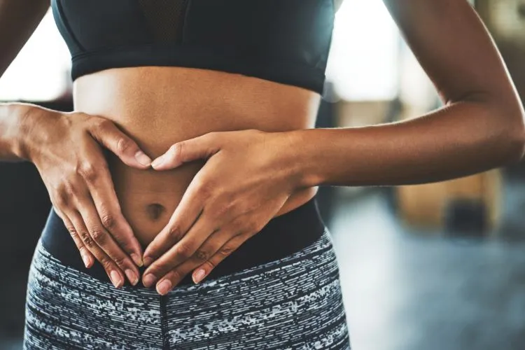 Mulher usando roupas esportivas formando um coração com as mãos sobre o abdômen, sugerindo satisfação com resultados obtidos possivelmente com o tratamento Mounjaro. Imagem simboliza saúde digestiva e bem-estar físico alcançado.