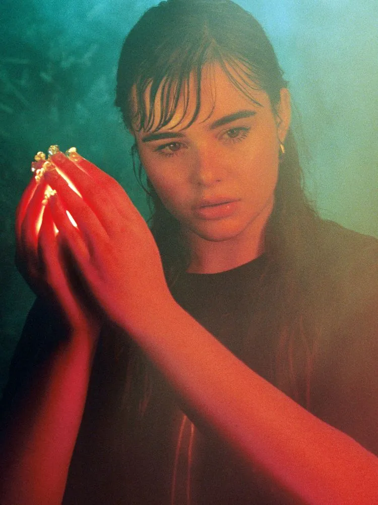uma foto de rosto da atriz Barbie Ferreira, uma luz está entre as mãos que ela tem levantadas em formato de concha