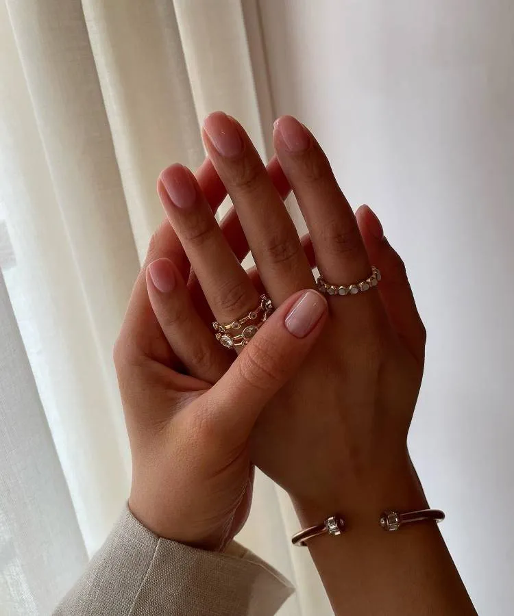 Mulher segura sua própria mão com a outra. A mão que ela segura possui anéis de pedras e um bracelete. Suas unhas elegantes são de cor rosa e possuem formato quadrado arredondado. As mãos estão em frente a uma parede com cortina.
