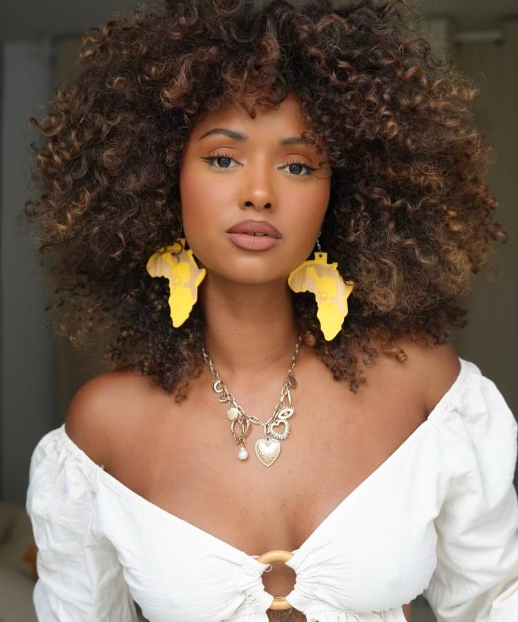 A imagem mostra uma mulher com cabelo castanho iluminado, longo e ondulado, que sorri amplamente. Ela usa um top branco e está em um ambiente externo com vegetação verde e luz solar ao fundo.








