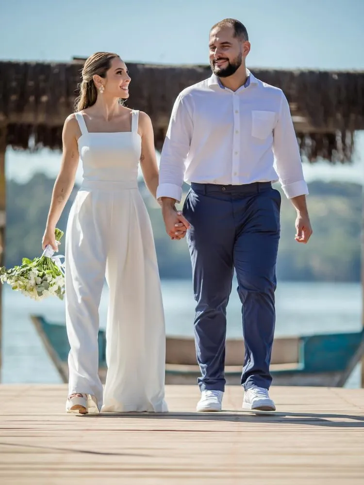 Mulher de pele clara de macacão branco ao lado de noivo com camisa branca e calça social azul
