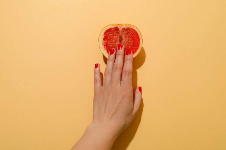 fundo amarelo com toranja cortada ao meio e mão de mulher com unhas vermelhas, desejo sexual feminino