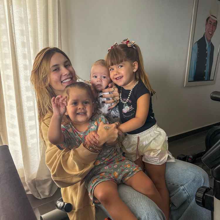 A imagem retrata Virginia Fonseca sorridente segurando três crianças (seus filhos) em um ambiente interno iluminado por uma cortina clara. As crianças estão felizes, incluindo um bebê nos braços. Ao fundo, há um retrato masculino emoldurado na parede.