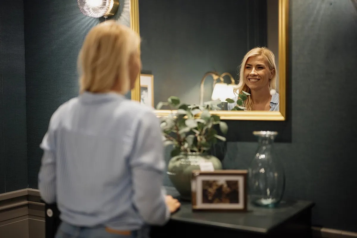Mulher loira de costas diante do espelho, sorrindo para o próprio reflexo