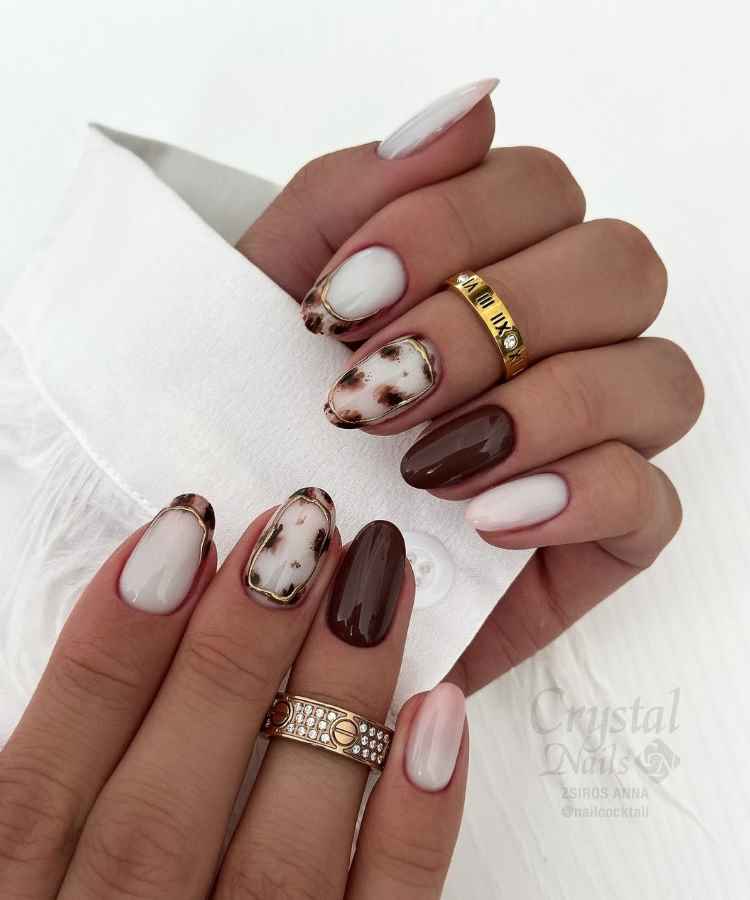 Unhas longas com esmalte em tons de branco, marrom e padrão tartaruga. Algumas têm bordas douradas elegantes. As mãos estão adornadas com anéis dourados e prateados, complementando o visual sofisticado. O fundo é claro, destacando o design das unhas.