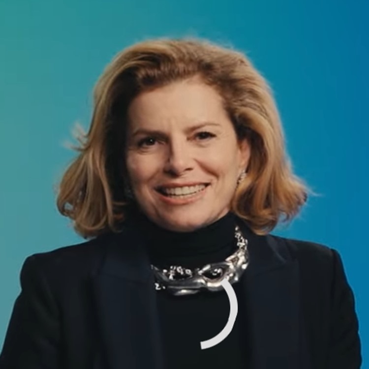 Mulher sorridente de cabelo loiro curto e volumoso, vestindo blazer escuro e gola alta preta com colar prateado chamativo. O fundo é azul suave, transmitindo um clima formal e acolhedor.





