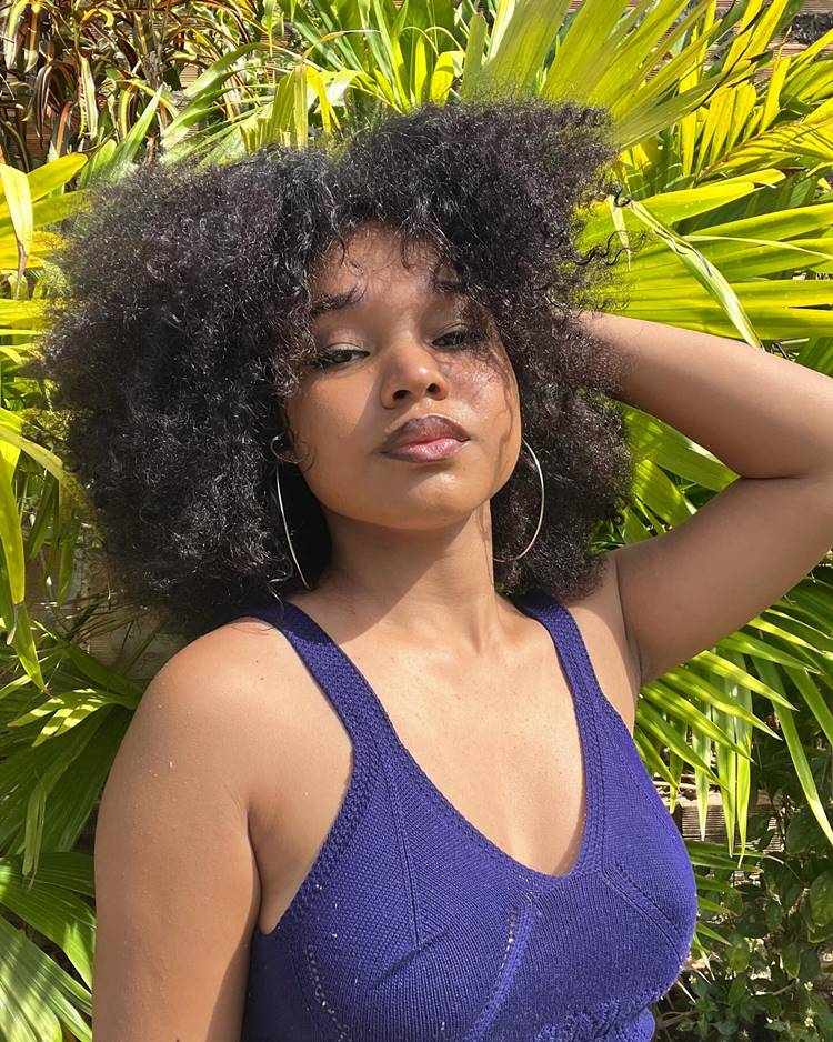 Menina com cabelo crespo e rosto redondo com corte blunt cut. Ela usa regata azul, brincos de argola e está em fundo com planta verde