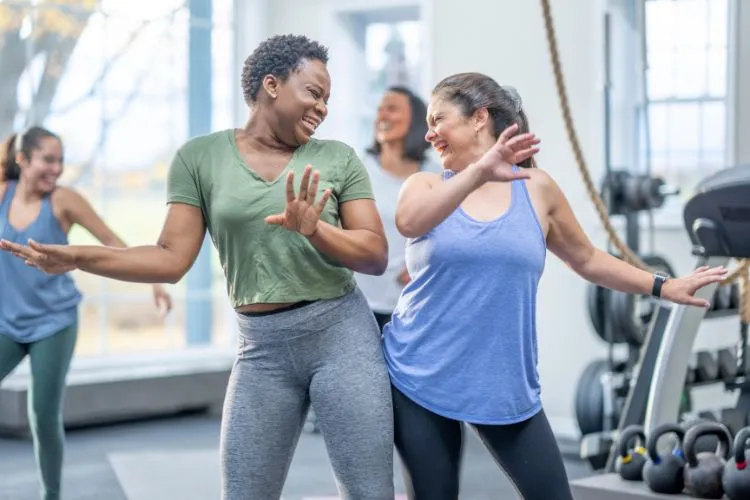 2 mulheres com os quadris encostados sorrindo e dançando em academia. Ao fundo, outras duas mulheres desfocadas sorrindo e dançando também