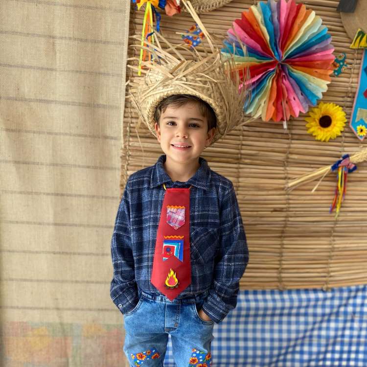 Menino usando camisa xadrez decorada, calça personalizada e chapéu de palha para festa junina.

