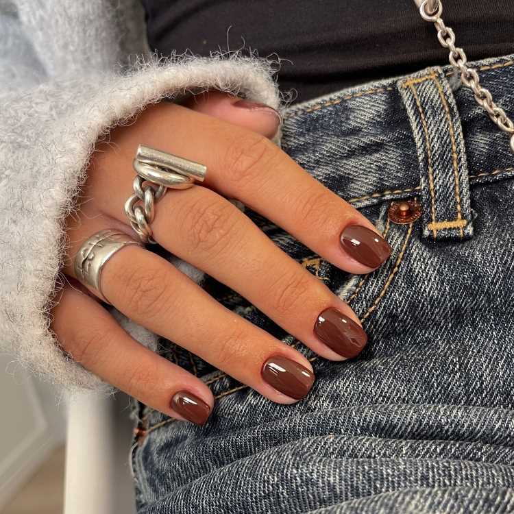 Close-up de uma mão com unhas pintadas de esmalte marrom brilhante, adornada com anéis de metal prateado. A mão repousa sobre jeans azul, e parte de uma blusa felpuda clara e corrente aparecem ao fundo.