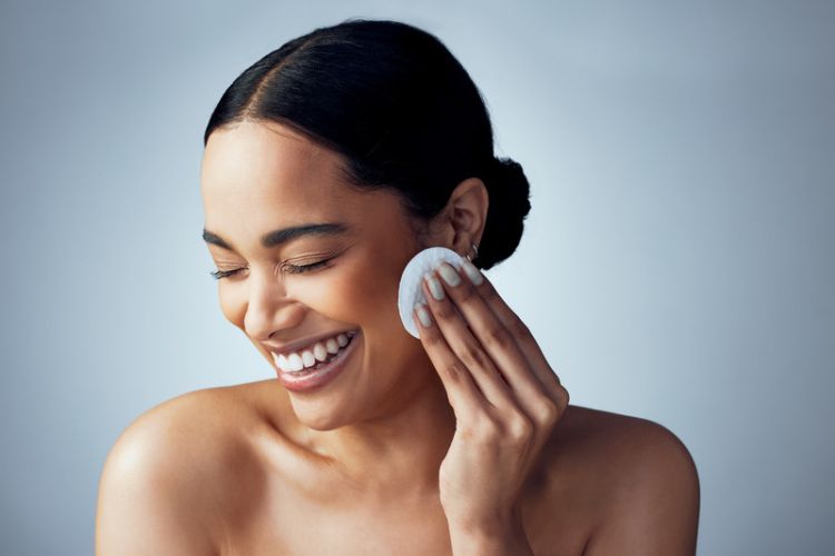mulher sorrindo e passando produto de cuidado com a pele no rosto usando um disco de algodão