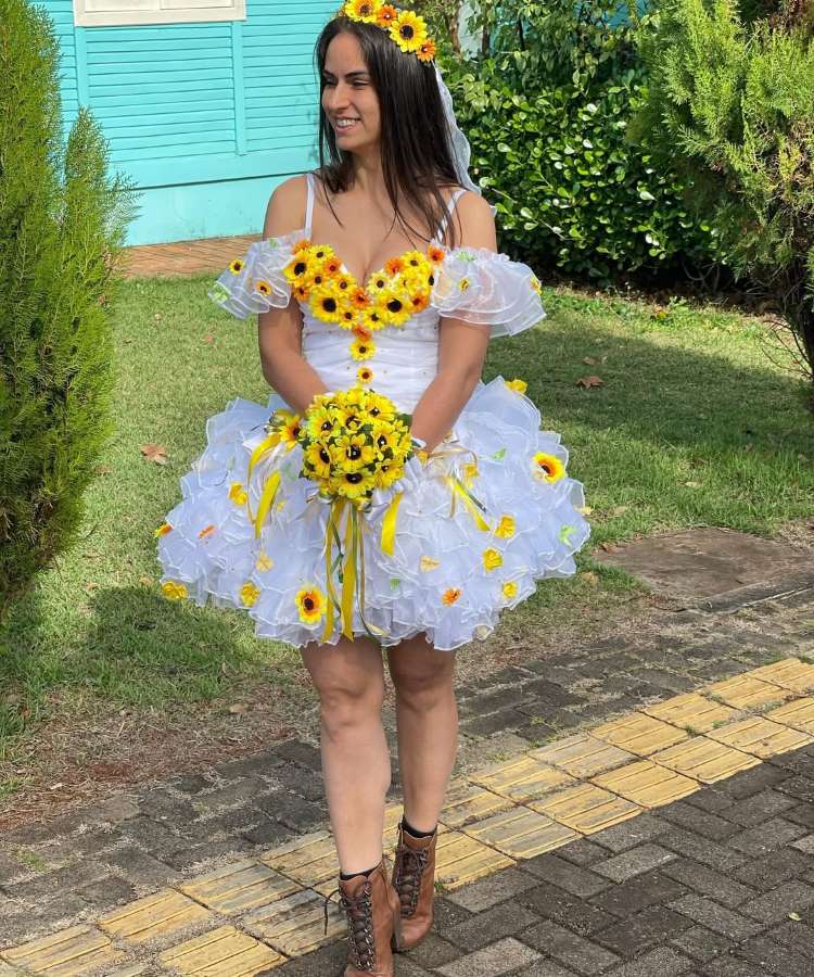 Uma foto vibrante ao ar livre de uma mulher com um vestido de noiva único decorado com girassóis e véu, segurando um buquê de girassóis. O sorriso e as cores vivas sugerem uma ocasião festiva como uma Festa Junina ou casamento temático, com vegetação exuberante ao fundo.