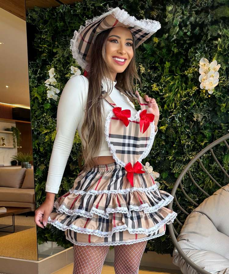 Uma mulher sorridente, com cabelos longos e castanhos, veste um traje junino xadrez bege e preto. Ela usa um chapéu de palha com renda, um top branco e uma saia de babados, além de meias arrastão brancas. O fundo é uma parede verde com orquídeas brancas.


Fontes

