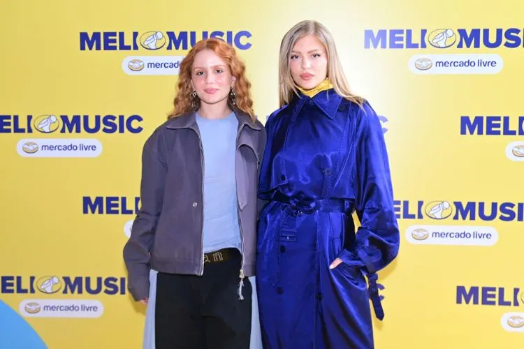 Carol Biazin e Luísa Sonza abraçadas em frente a um painel do Meli Music 