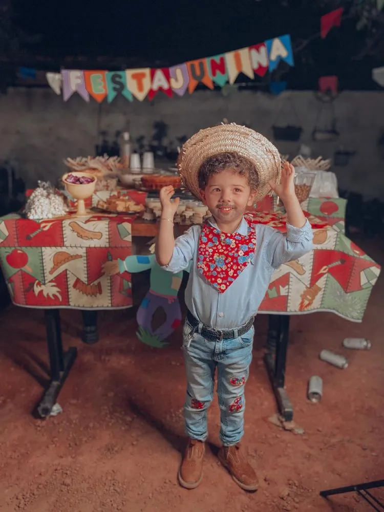 Menino de pele clara em festa junina usando camisa, lenço, chapéu de palha, bota e calça jeans com retalhos 