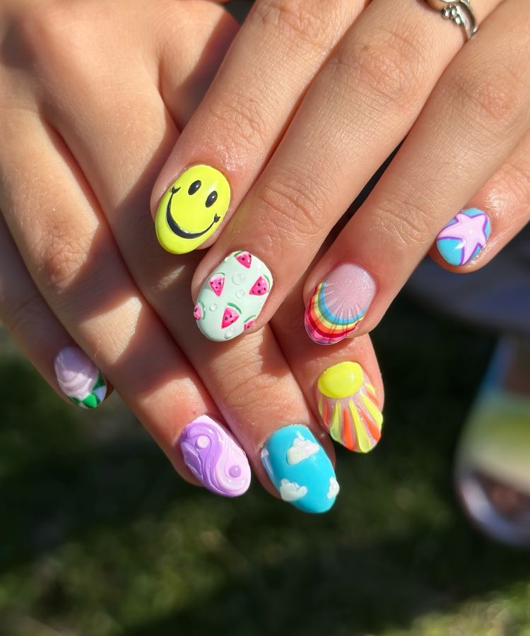 A imagem exibe unhas de carnaval vibrantes e divertidas. Cada unha apresenta um design único, com temas como sorriso amarelo, melancias, arco-íris, estrelas e nuvens. As cores vivas, como amarelo, rosa e azul, criam um visual alegre e criativo.