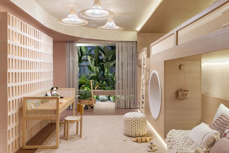 A imagem mostra um quarto infantil bem iluminado com uma grande janela aberta para a vegetação externa. Há uma cama embutida, uma escrivaninha de madeira, brinquedos no chão e decoração suave.

