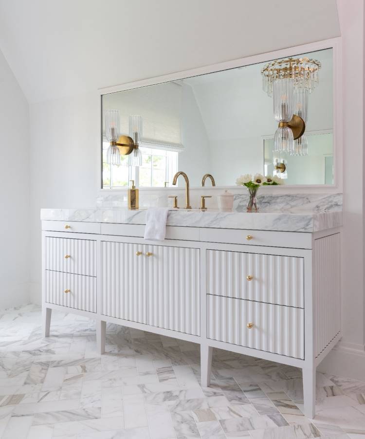A imagem apresenta um banheiro sofisticado com gabinete branco de ripas e puxadores dourados. A pia de mármore possui torneiras douradas elegantes. Um grande espelho reflete a luminária de cristal pendente. O piso é de mármore em tons claros.