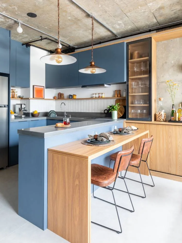 A imagem mostra uma cozinha moderna com um design que combina diferentes materiais. Há concreto visível em parte das paredes e no teto, contrastando com elementos de madeira usados predominantemente nos armários e na ilha central. A ilha possui uma bancada que se estende, criando uma área de bar com três bancos altos de pernas de metal preto e assentos de couro marrom. Acima da ilha, pendem duas luminárias com acabamento em latão, suspensas do teto de concreto exposto.

A parede ao fundo da cozinha é equipada com bancadas e armários cinza escuro, acompanhados por um backsplash em tons de cinza mais claros. Há também prateleiras embutidas cheias de copos ao lado de uma geladeira que se integra aos armários. Na bancada da ilha, há pratos dispostos como se estivessem prontos para uma refeição, junto com alguns itens decorativos como vasos.