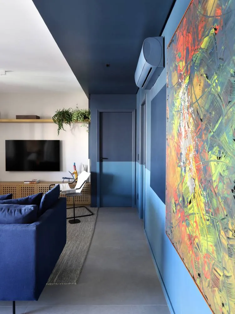 Esta imagem mostra uma sala de estar moderna com um sofá azul e uma grande TV. Um corredor adjacente exibe paredes azuis com um teto colorido em azul-marinho. Uma porta discreta se mistura à parede, e uma obra de arte abstrata vibrante adiciona um toque de cor.

