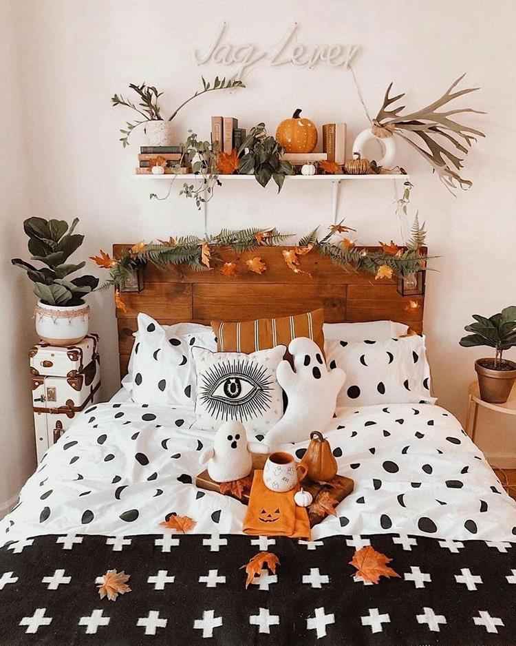 quarto com decoração de halloween, com abóboras em prateleira, galhos secos, flores laranjas, fantasmas de pelúcia, roupa de cama em preto e branco e mais