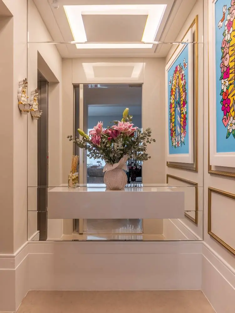 Hall de entrada com espelho, aparador branco, vaso com flores rosas, parede off white com boiserie dourado e quadro colorido 