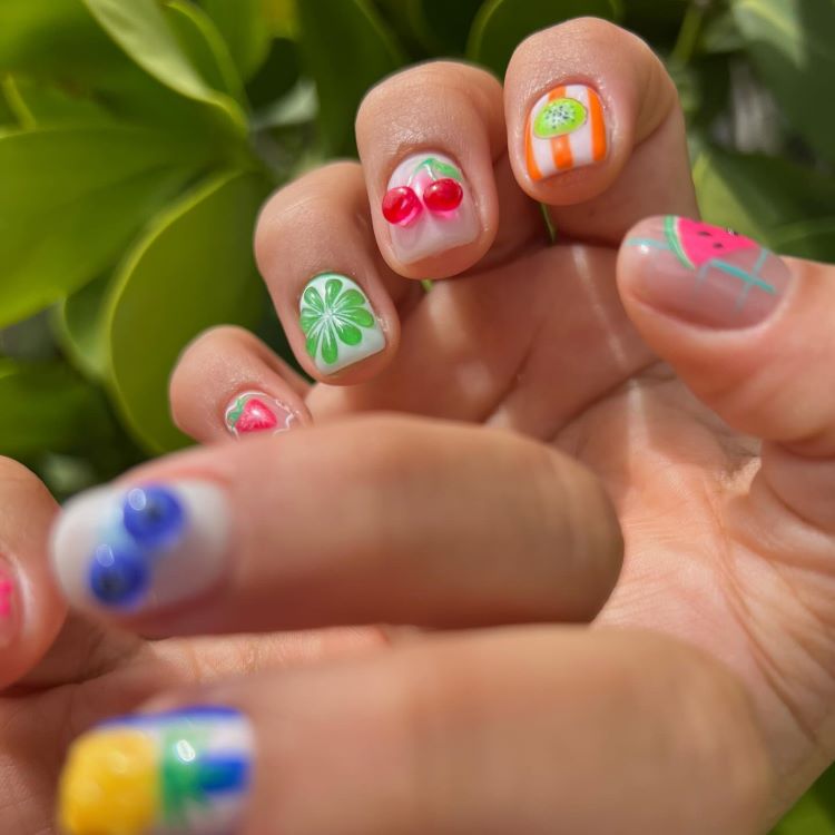 Unhas decoradas com frutas e desenhos coloridos: cerejas, kiwi, melancia, trevo verde, abacaxi e mirtilos. Base clara destaca os detalhes vibrantes. Mãos iluminadas pelo sol, em frente a folhas verdes desfocadas, criando um visual alegre e tropical.